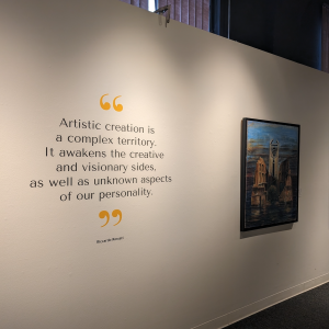 One of the white walls of the exhibition with a quote from the artist and a painting in the right corner. The quote reads Artistic creation is a complex territory. It awakens the creative and visionary sides, as well as unknown aspects of our personality. The picture is taken in perspective.