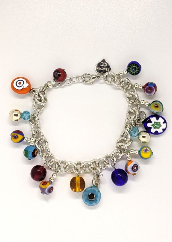A charm bracelet is being crafted using the traditional millefiori (thousand-flowers) technique, where thin rods of glass (called murrine) are carefully arranged in a pattern and then heated until they fuse together. The bracelet is designed in bright colors reminiscent of carnevale.