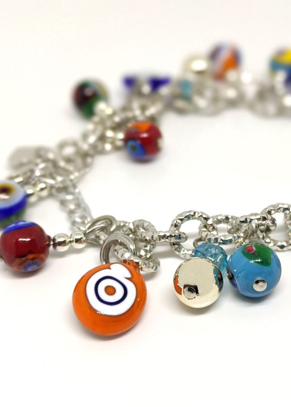 A charm bracelet is being crafted using the traditional millefiori (thousand-flowers) technique, where thin rods of glass (called murrine) are carefully arranged in a pattern and then heated until they fuse together. The bracelet is designed in bright colors reminiscent of carnevale.