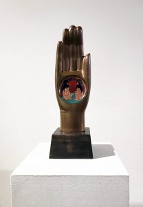 A hand sculpted from bronze and enamel, placed upon a wooden base. Each of the fingers are straight and together, touching one another. Within the open palm is a circle which contains four young children; two in the foreground, sitting unclothed with their knees close to their chest upon green ground, mirroring one another, and two in the background, facing one another. The hand itself is bronze in color, while within the circle is a background with a sky of red, purple, and orange rings.